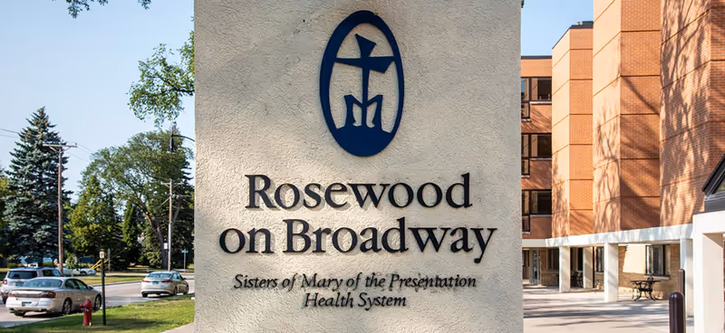 Outdoor view of the entrance sign for Rosewood on Broadway, part of the Sisters of Mary of the Presentation Health System, with a multi-story brick building and trees in the background.