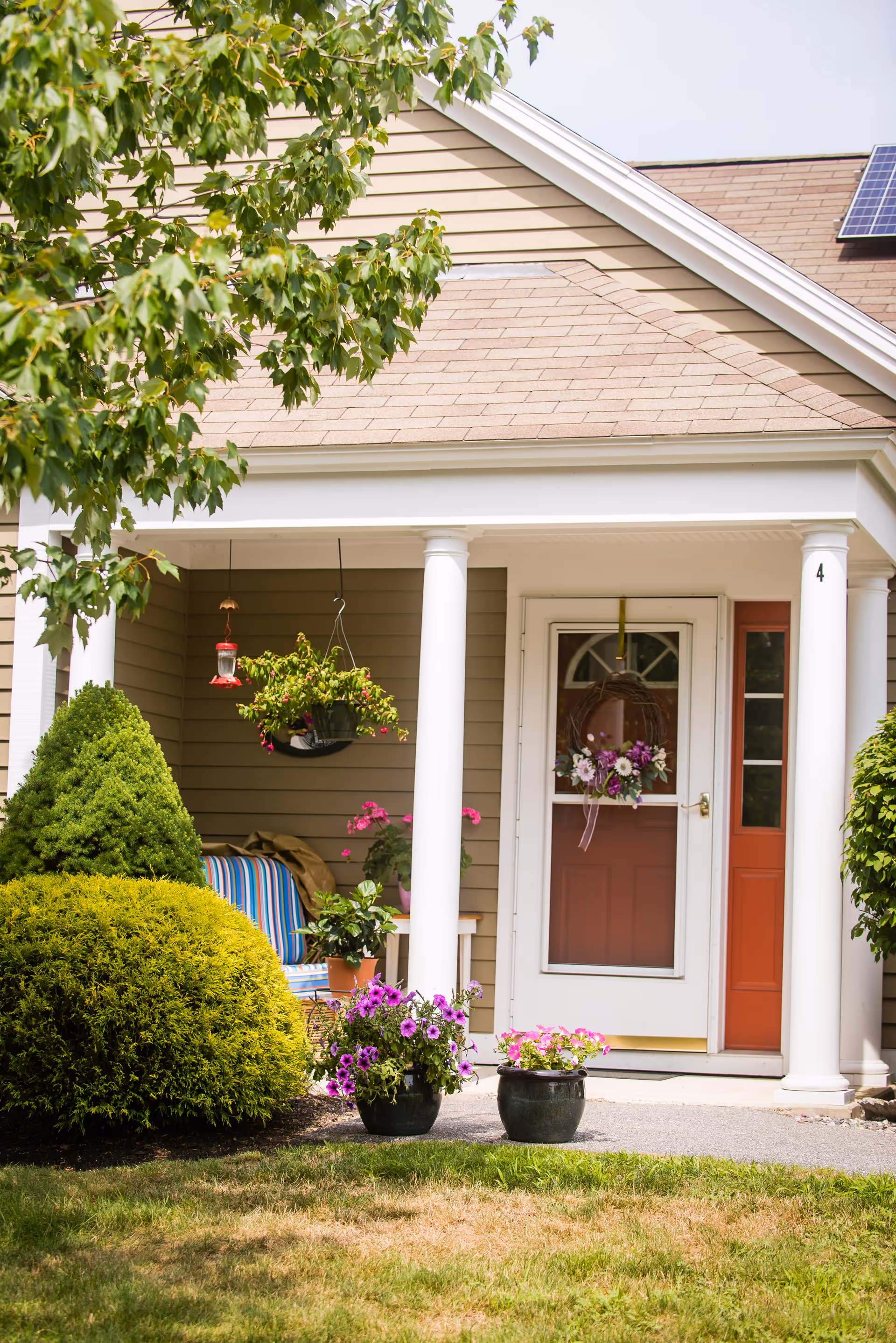 Front porch of a house with beige siding and a brown roof. The porch has white columns and a red door decorated with a floral wreath. There are hanging flower pots and potted plants with purple and pink flowers on the porch and in front of it. A striped chair is partially visible on the porch. Green bushes and a tree branch frame the scene.
