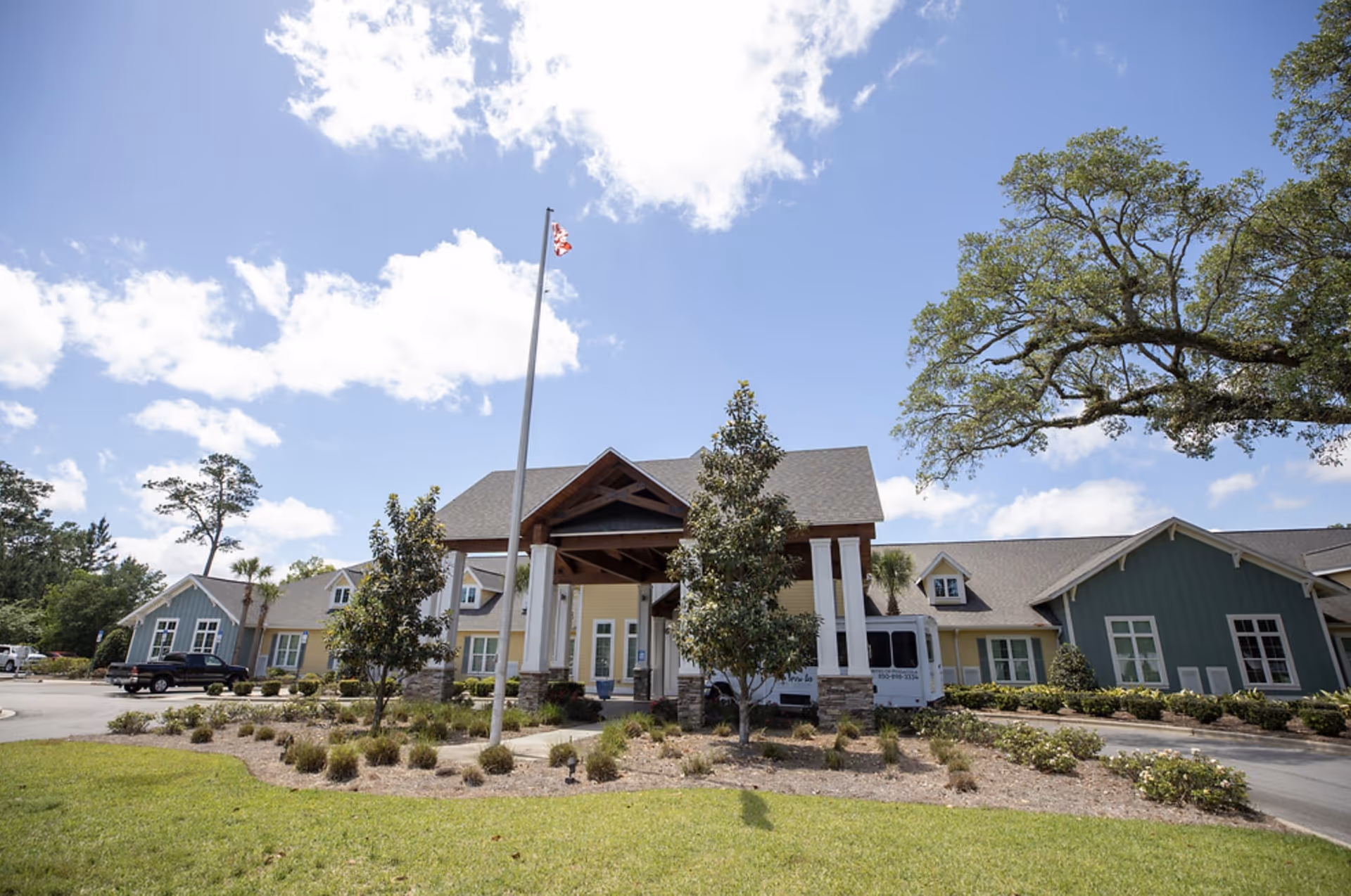 Front exterior of a single-story senior living facility with a covered entrance, flagpole, landscaped grounds, and a bright blue sky.
