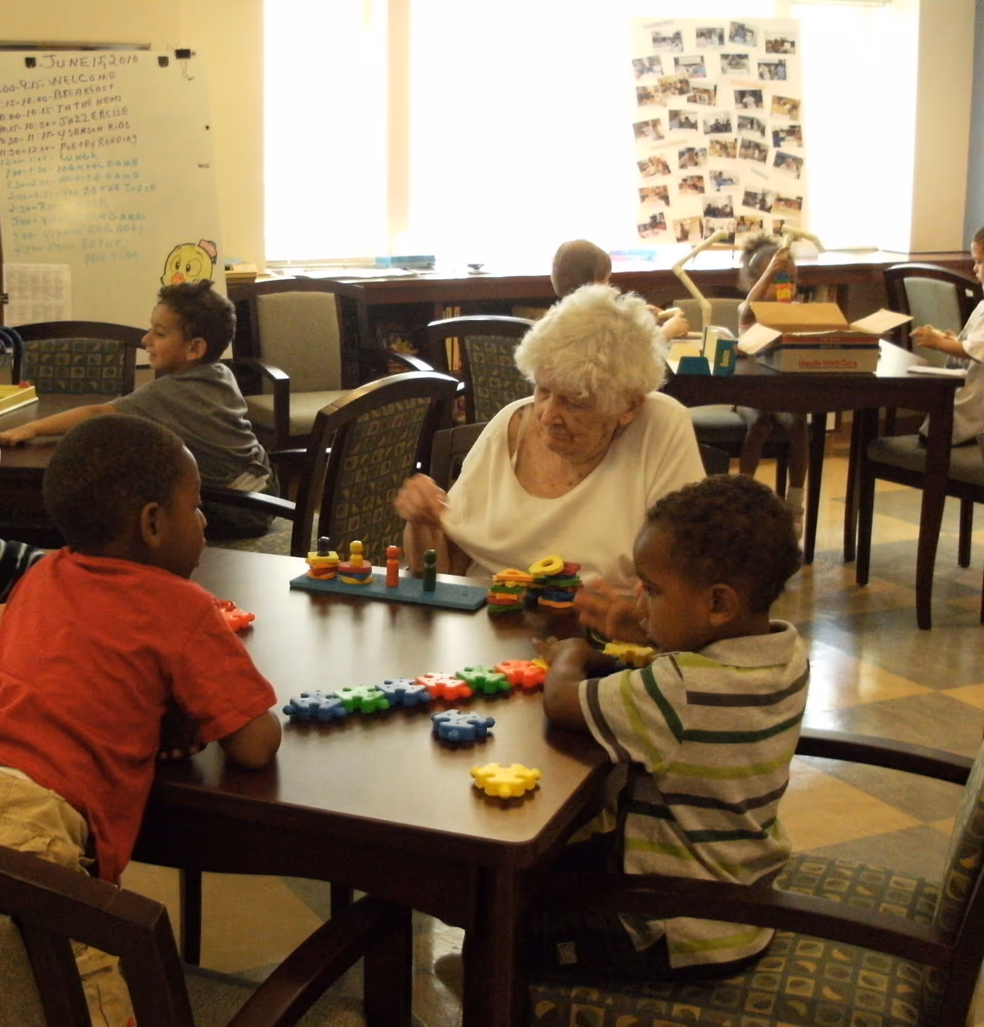 An elderly woman and several young children sit around tables in a well-lit room, engaging in activities with colorful educational toys. The room has chairs with patterned upholstery, a whiteboard with a schedule, and a bulletin board with photos on the wall.