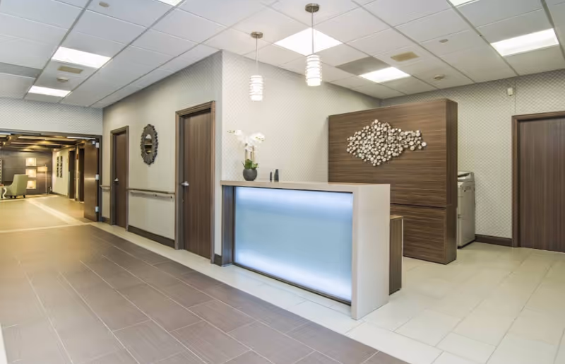Reception area in Alden Care Forest Park featuring a modern front desk with a glowing blue panel, decorative wall art, and three wooden doors along the hallway. The space has tiled floors, a drop ceiling with recessed lighting, and pendant lights above the desk. A hallway leads to a lounge area with seating and lamps.