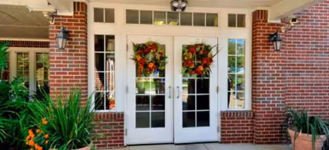 Brick building exterior with white double doors, wreaths, and potted plants at entrance.