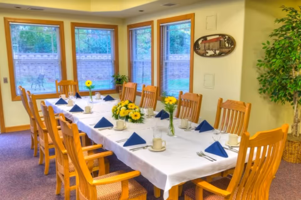 A dining room with a long rectangular table covered with a white tablecloth. The table is set with blue folded napkins, cups, glasses, and silverware. There are yellow flower centerpieces on the table. Wooden chairs surround the table. Three large windows with blinds let in natural light, and a green potted plant is in the corner of the room.