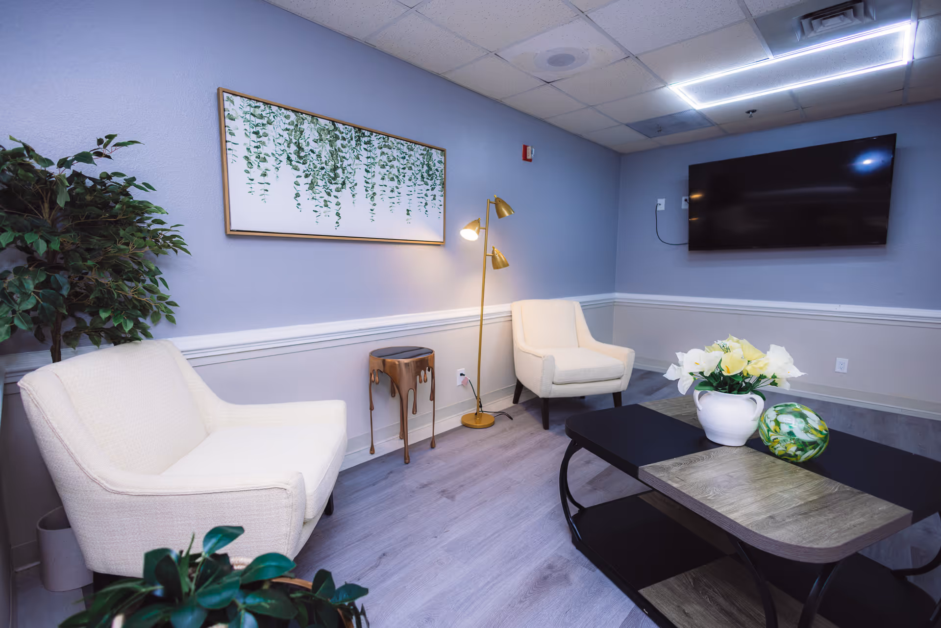 A cozy sitting area with two white armchairs, a black and wood coffee table with a white vase of flowers and a decorative green glass orb, a tall gold floor lamp, a small artistic side table, a large flat-screen TV mounted on a light purple wall, and a framed artwork of hanging green leaves. There are also two potted plants in the room and light wood flooring.