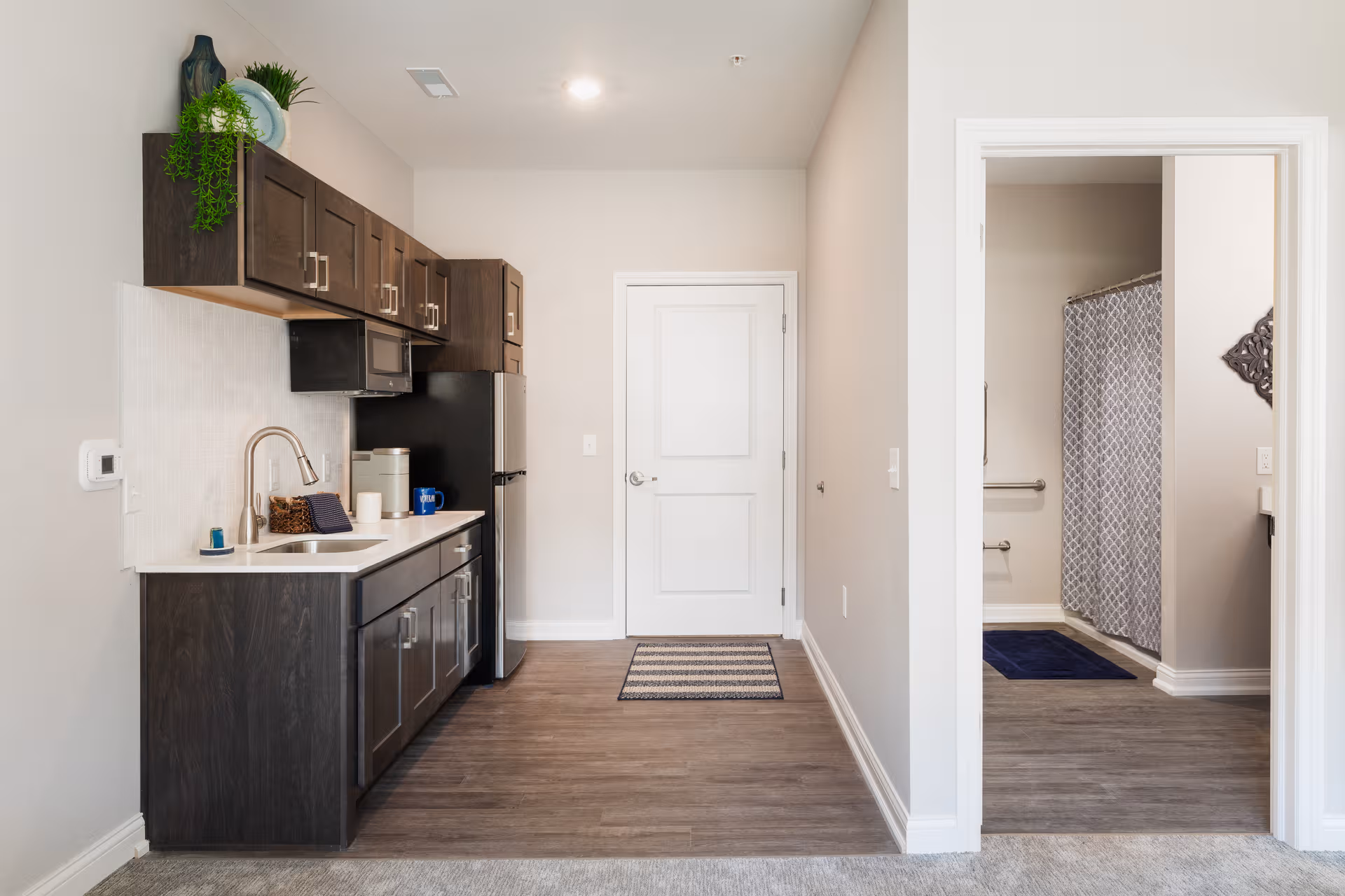 Modern kitchenette with dark cabinets, sink, microwave, fridge, and doorway to bathroom.