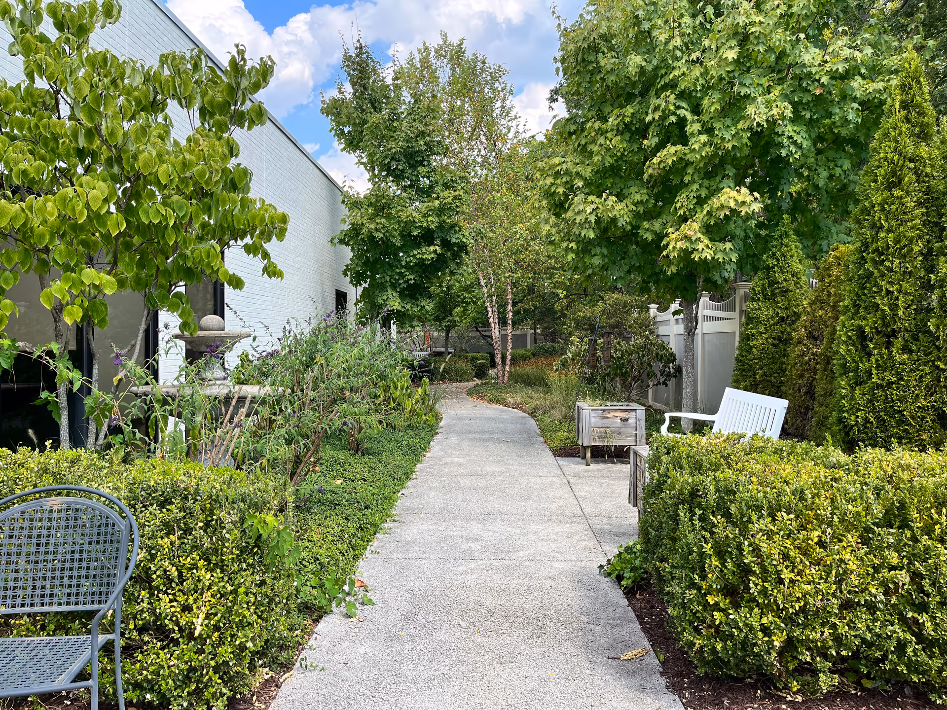A paved garden pathway lined with green bushes and trees on both sides. There is a white bench and a small wooden planter box on the right side, and a metal chair partially visible on the left. The sky is partly cloudy above.