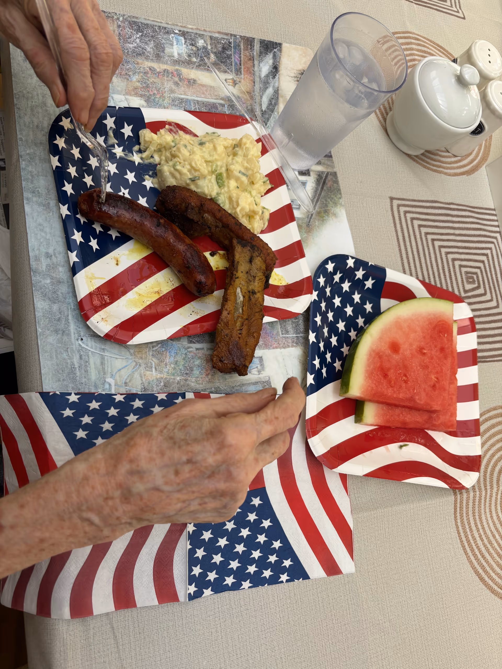 Elderly hands at a dining table with American-flag plates, sausages, and watermelon slices.