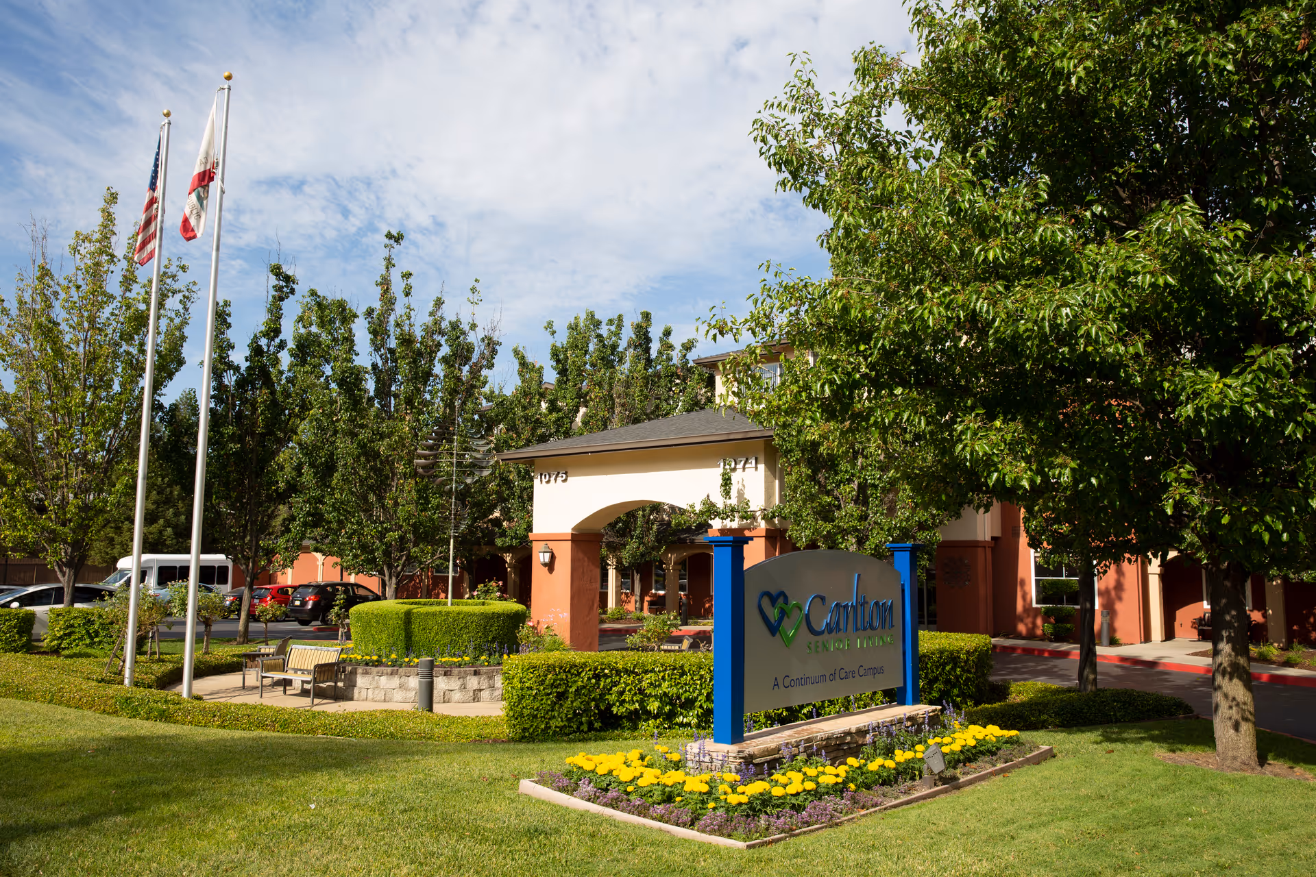 Exterior view of Carlton Senior Living Sacramento with sign, landscaping, and flagpoles.