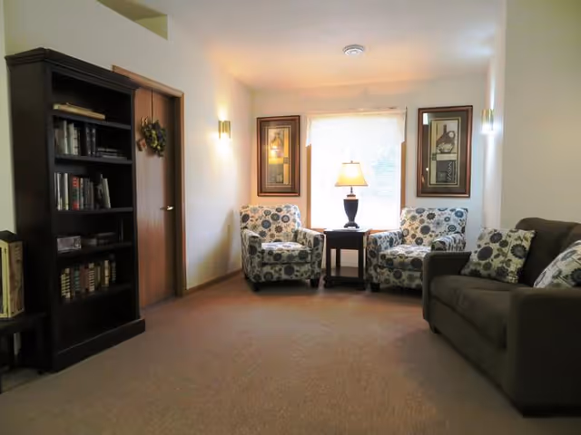A cozy living room area with two patterned armchairs and a dark brown sofa with matching pillows. Between the armchairs is a small wooden table with a lamp on it. The room has beige walls, a window with a sheer curtain, two framed pictures on either side of the window, a wooden bookshelf filled with books, and a wooden door with a small wreath hanging on it.