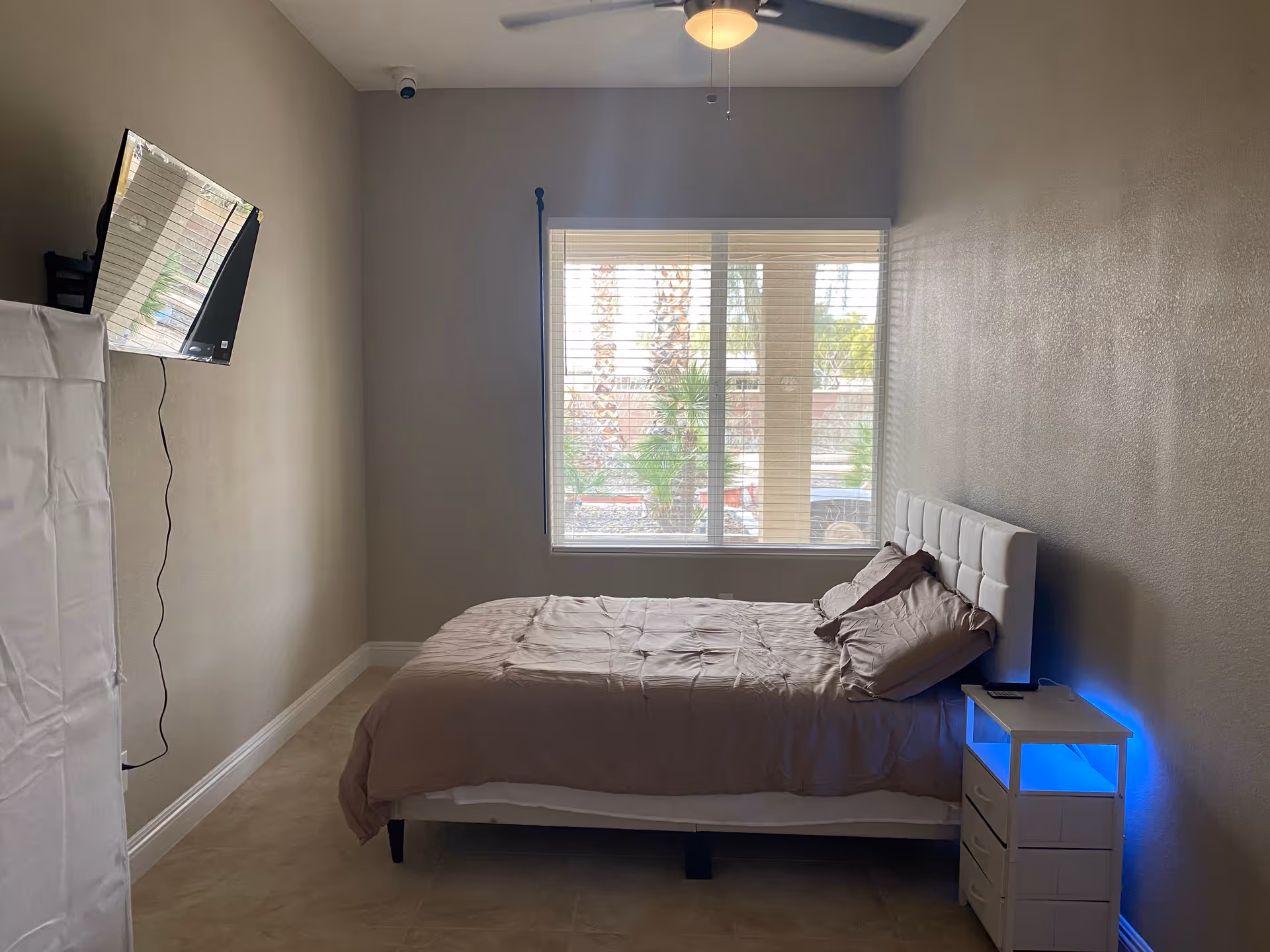 A compact bedroom with a bed, bedside table, ceiling fan, wall-mounted TV, and window.