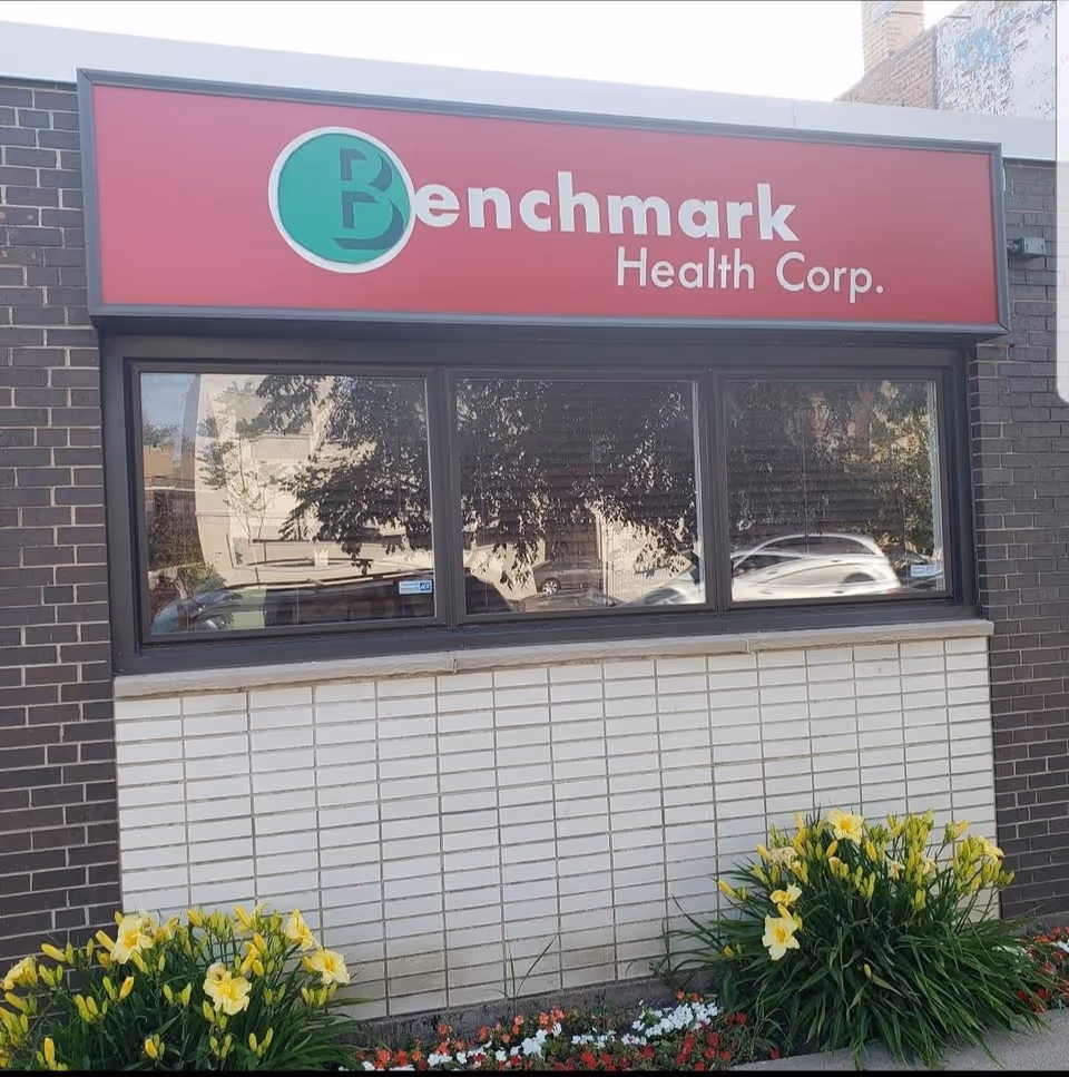 Red Benchmark Health Corp sign above a storefront window on a brick building with flowers.