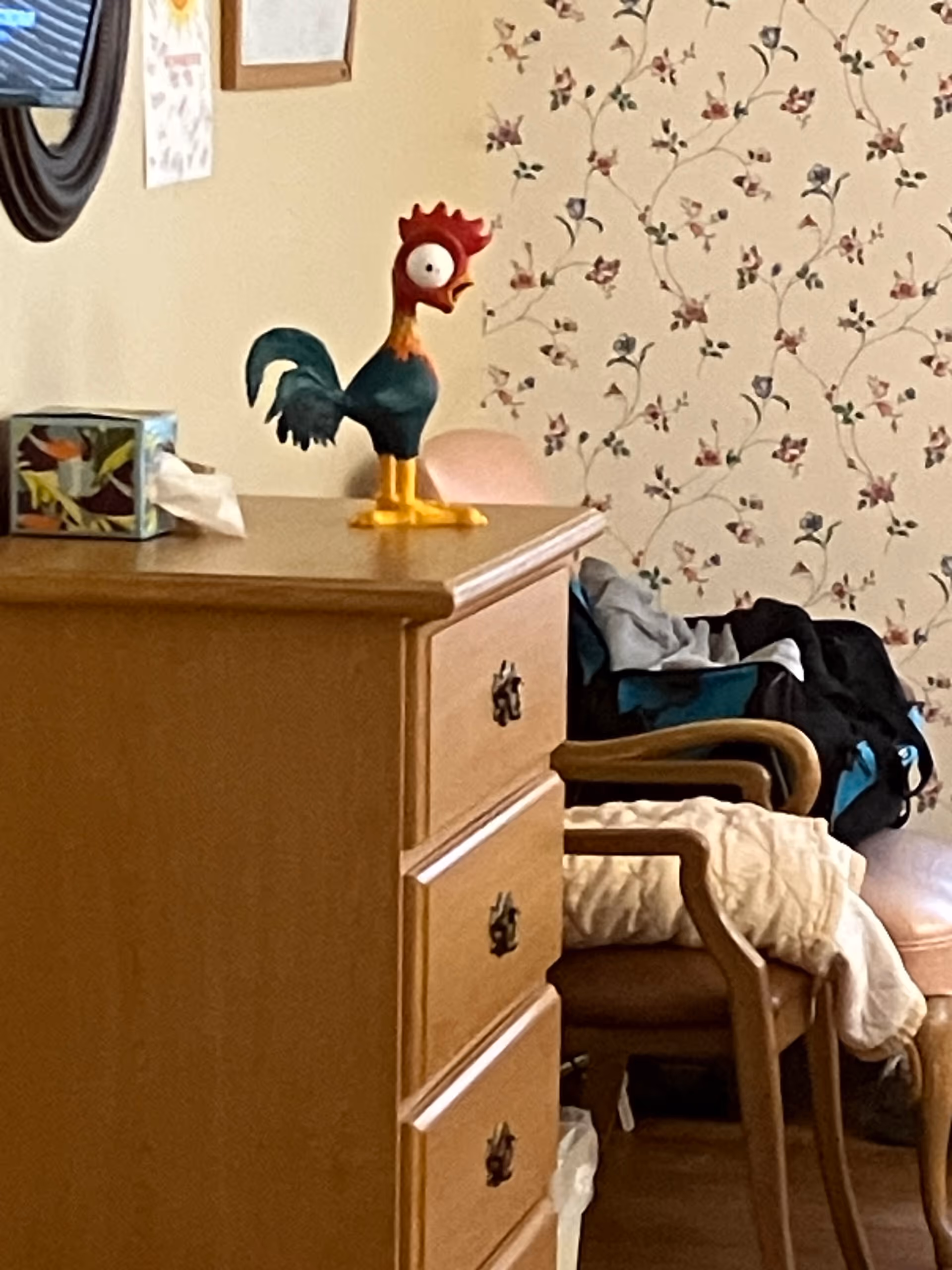 Wood dresser with a rooster statue, floral wallpaper, and a chair in a cluttered room.