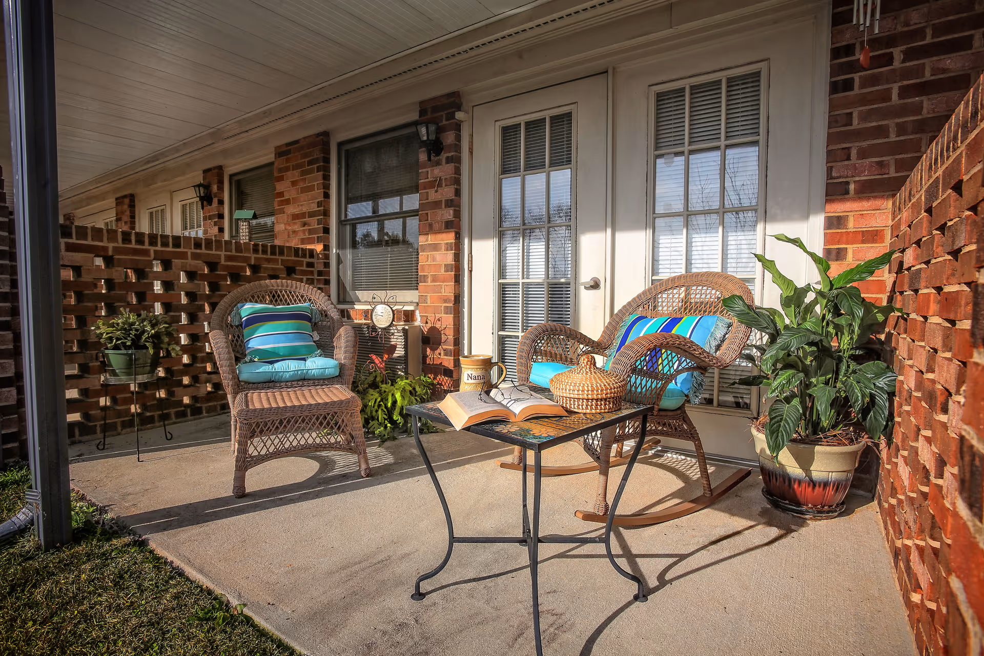 A cozy outdoor patio area with two wicker chairs featuring blue and green striped cushions, a small metal table with an open book, a woven basket, and a mug labeled 'Nana'. The patio is surrounded by brick walls and has potted plants, with a set of white French doors and a window in the background.