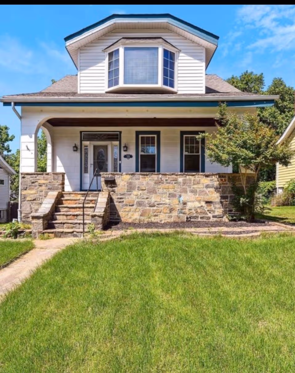 Front view of a stone-porch house with green lawn and bright blue sky.