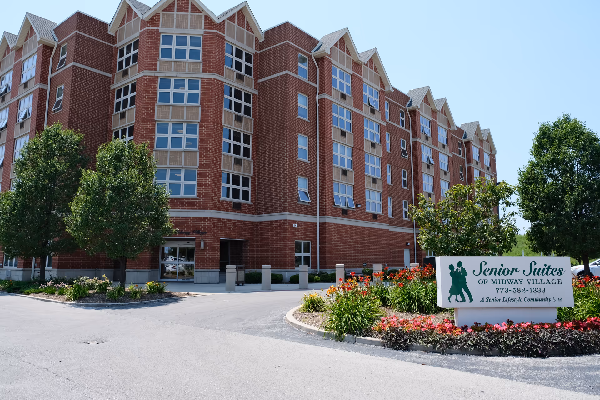 Brick multi-story senior living building with landscaped entrance and a sign reading 'Senior Suites of Midway Village'.