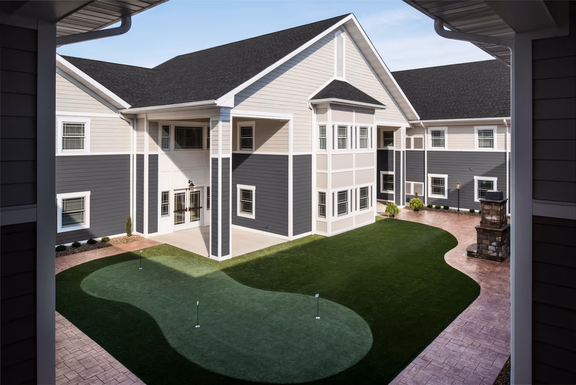 View of a courtyard at The Inn at Poland Way featuring a small artificial putting green with three holes, surrounded by a paved walkway and a two-story building with gray and white siding and multiple windows.