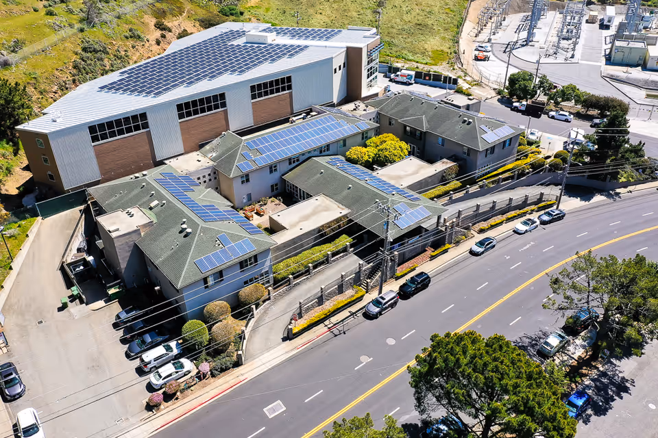 Aerial view of Mission Villa Senior Living facility showing multiple buildings with solar panels on the roofs, surrounded by roads, parked cars, and greenery.