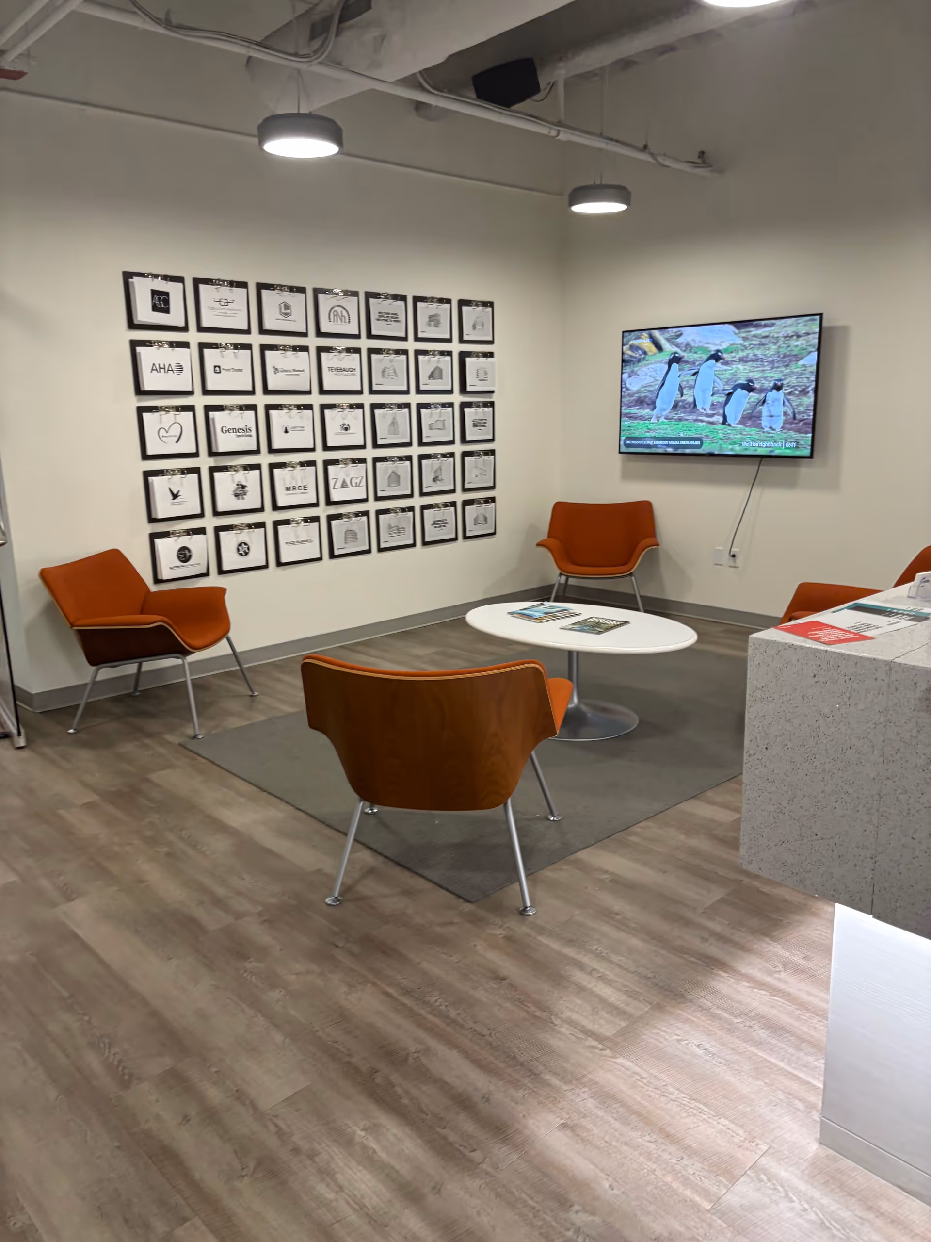 Modern waiting lounge with orange chairs, framed certificates, and a wall-mounted TV.