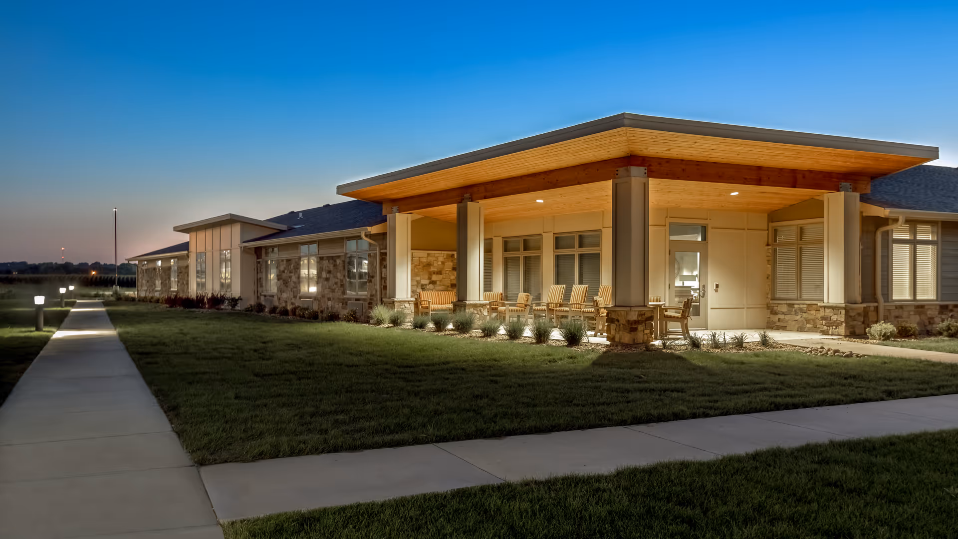 Exterior view of Brookestone Gardens facility at dusk with a well-lit covered porch area featuring wooden rocking chairs, surrounded by a neatly maintained lawn and a paved walkway with small lights.