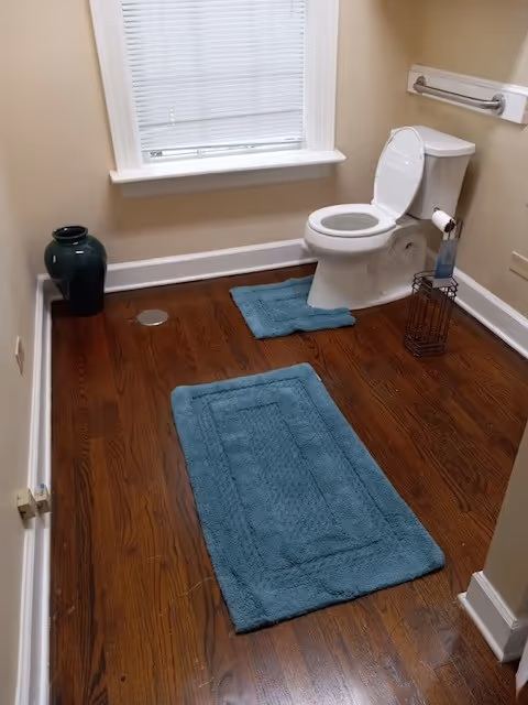 Bathroom with toilet, blue rugs, window, and wooden floor.