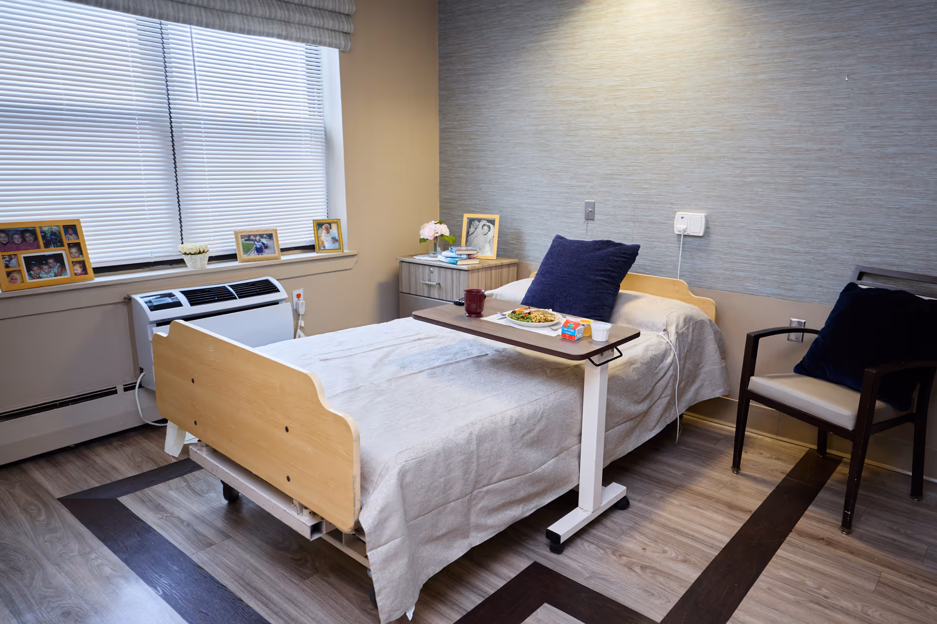 A senior care patient bedroom with bed, tray, chair, and photos near a window.