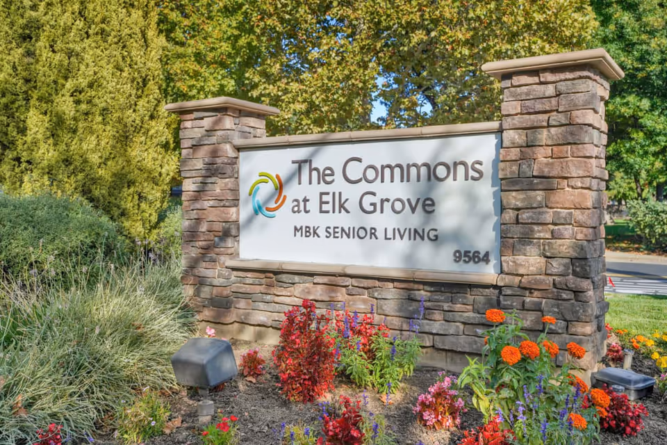 Stone sign for The Commons at Elk Grove MBK Senior Living surrounded by colorful flowers and greenery with trees in the background.