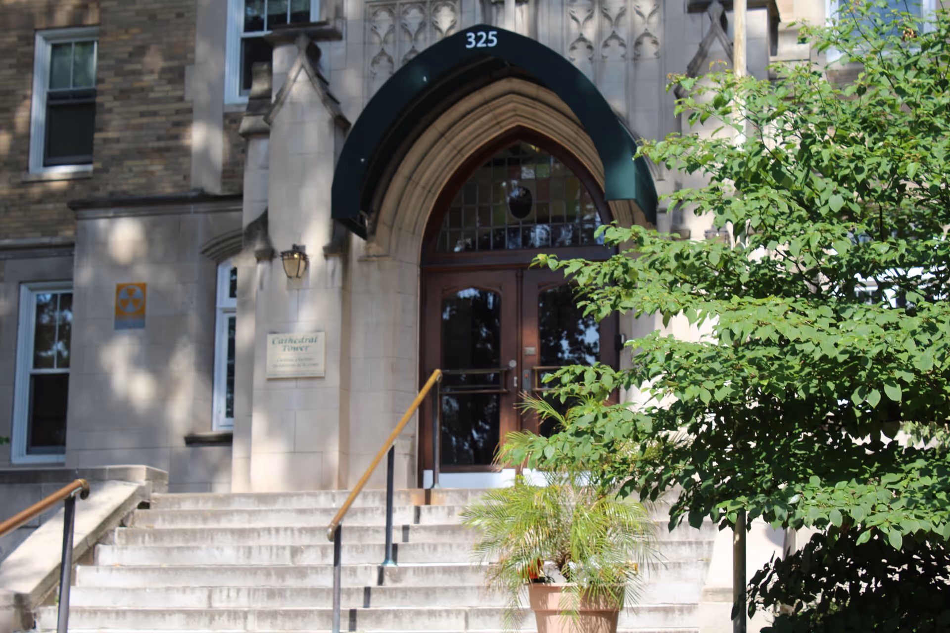 Stone building entrance with steps leading to arched double doors under a green awning numbered 325, flanked by a potted plant and leafy tree.