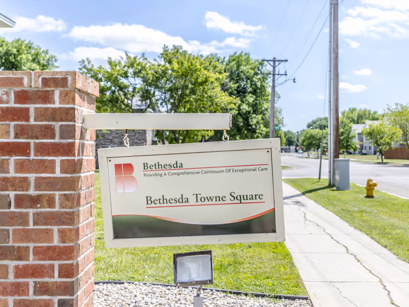 A hanging sign reading "Bethesda Towne Square" mounted on a brick post beside a sidewalk and street.