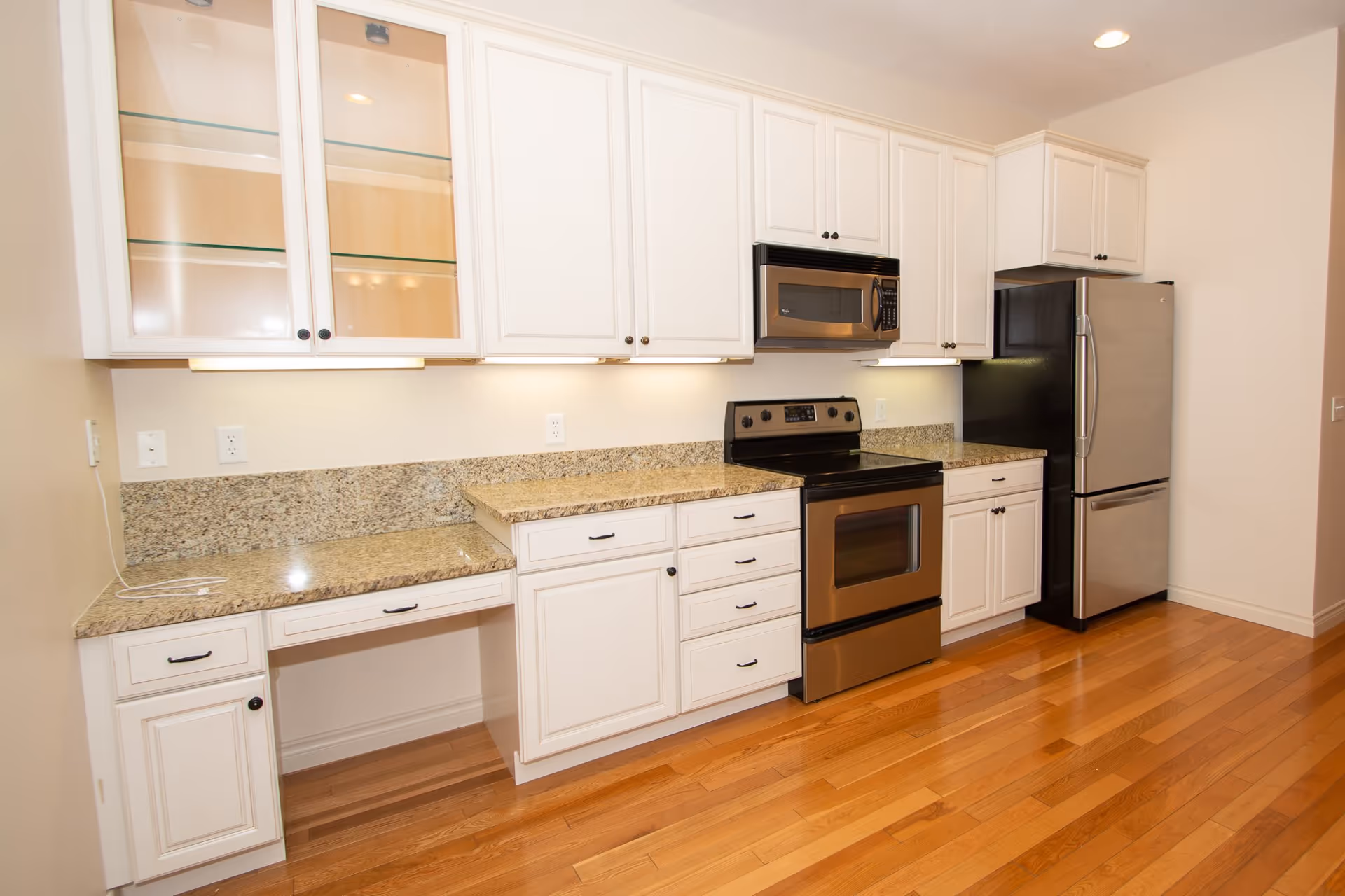 A modern kitchen with white cabinets, granite countertops, a stainless steel refrigerator, stove, and microwave. The floor is wooden and the kitchen is well-lit with under-cabinet lighting.