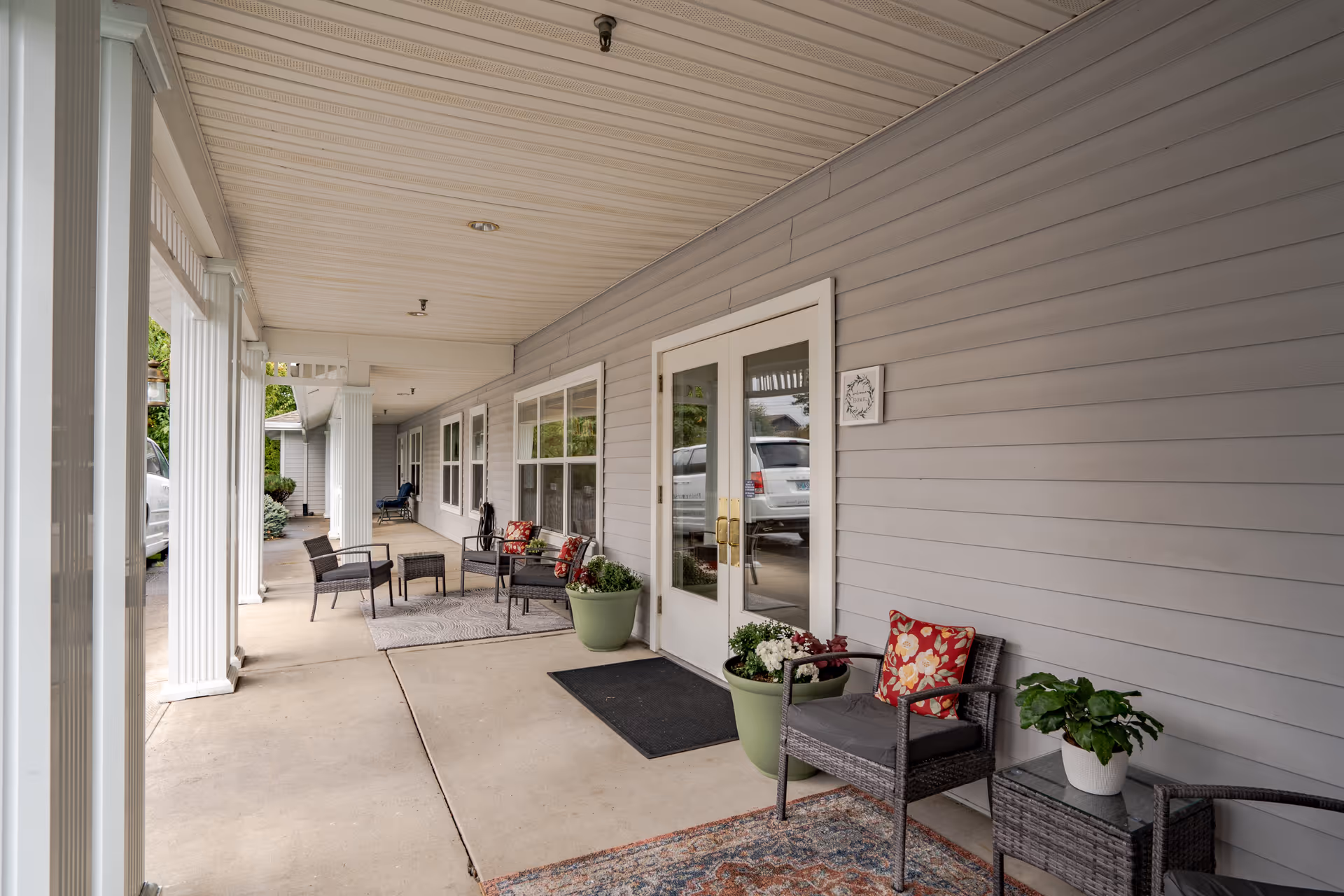 Covered front porch with wicker chairs, potted plants, rugs, and double glass entry doors.