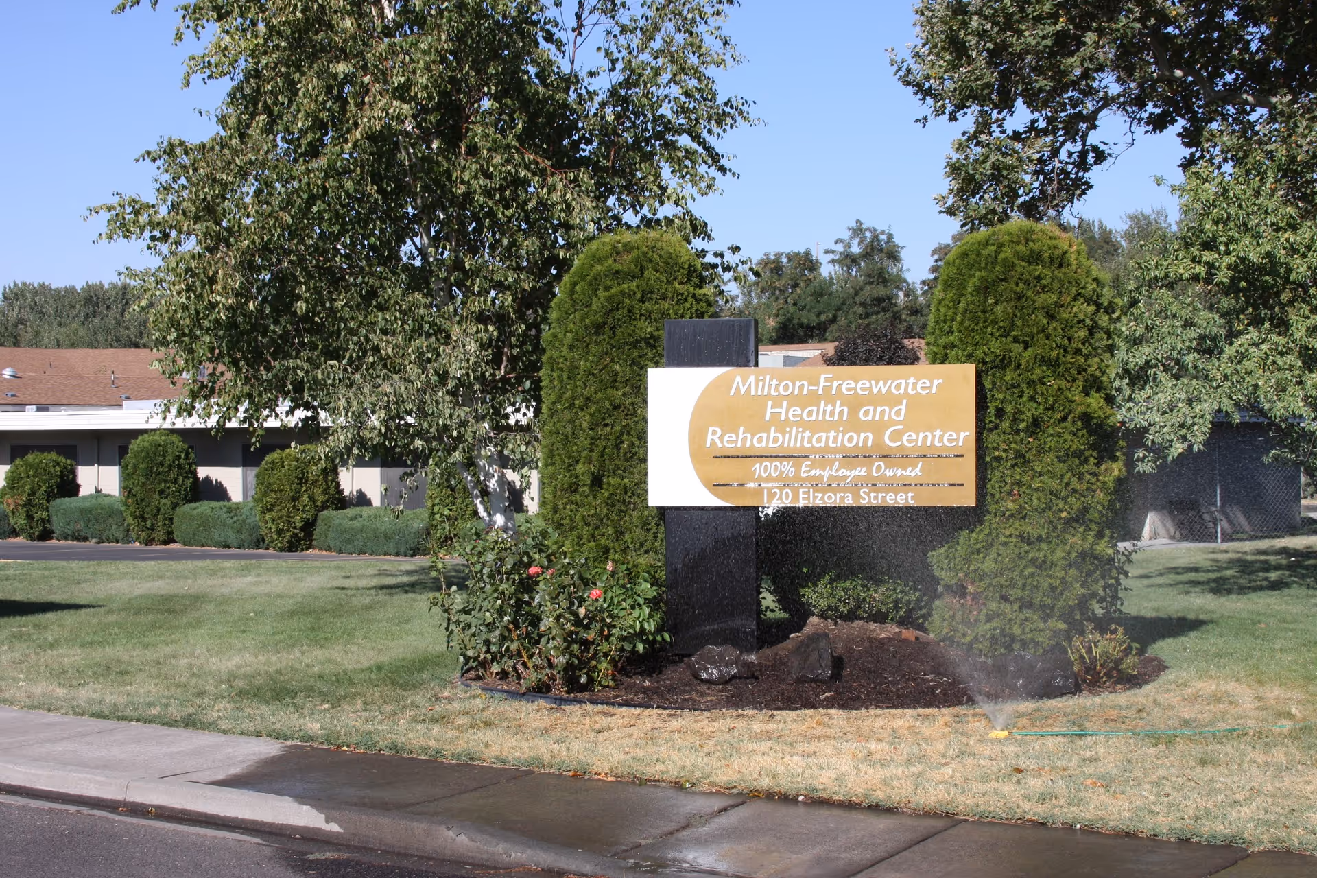 A landscaped lawn and trees with a prominent sign reading "Milton-Freewater Health and Rehabilitation Center" in front of the facility.