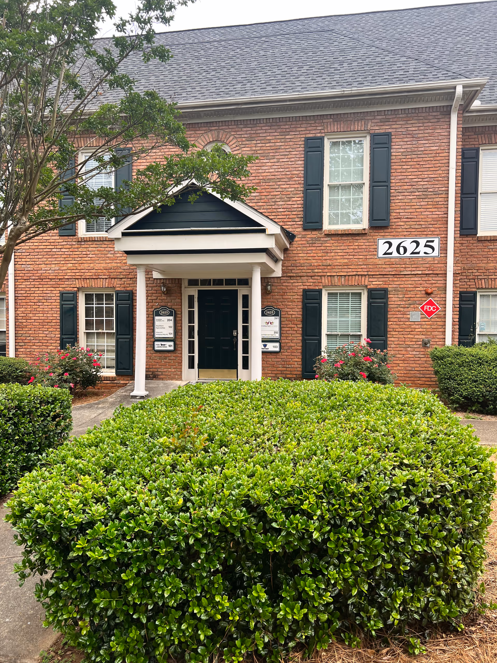 Brick senior living building exterior with shutters, greenery, and a covered entrance.