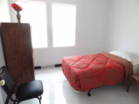 A simple bedroom with a single bed covered in a red patterned comforter, a wooden dresser with a vase of red flowers on top, a black chair, and two large windows letting in natural light.