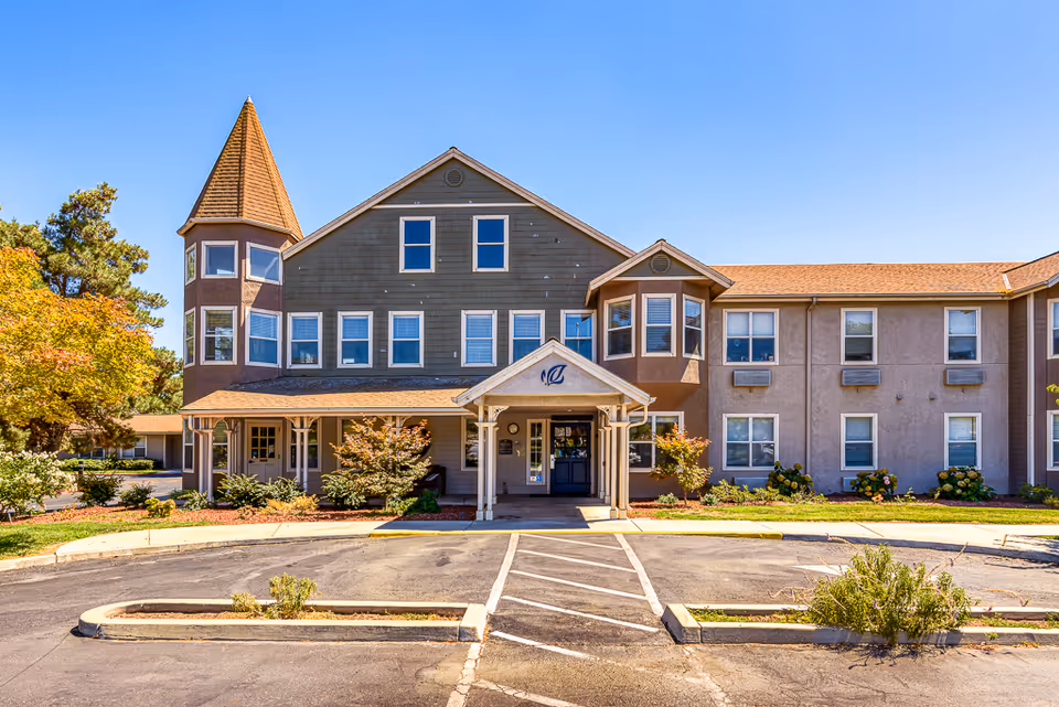 Front exterior of a two-story senior living facility with a turret, covered entrance, landscaping and parking area.