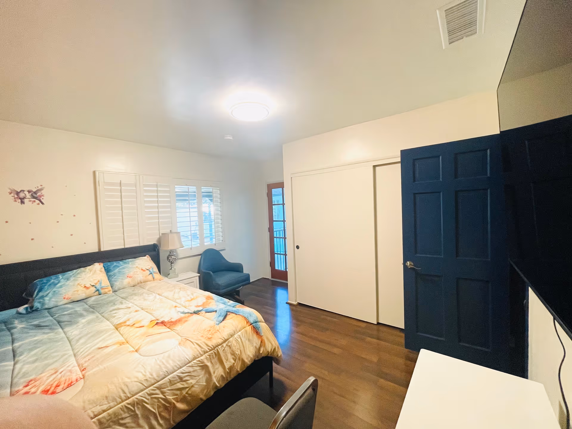 A furnished bedroom with a bed, nightstand, chair, and blue door in Broadmoor Assisted Living.
