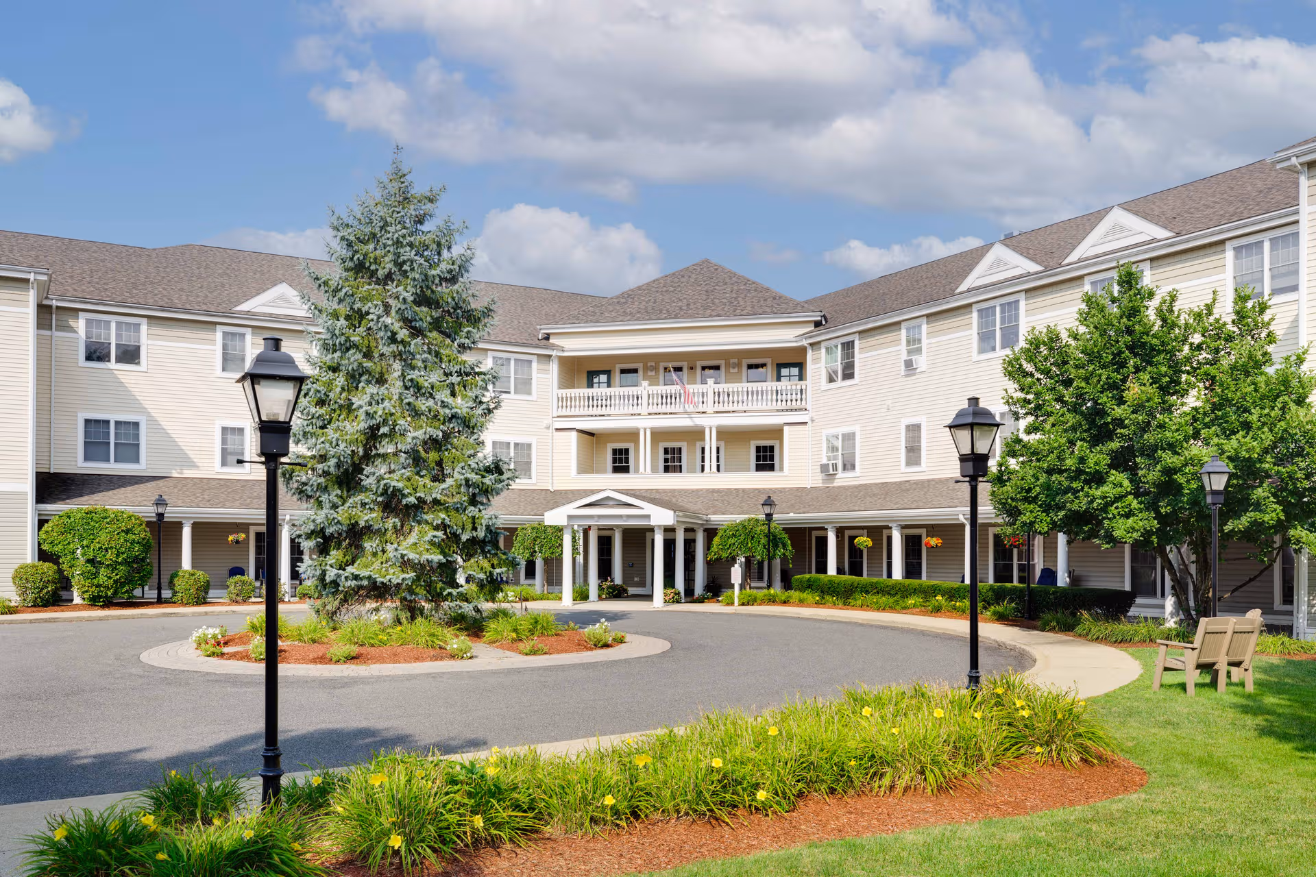 Senior living facility exterior with circular driveway, landscaping, and benches.