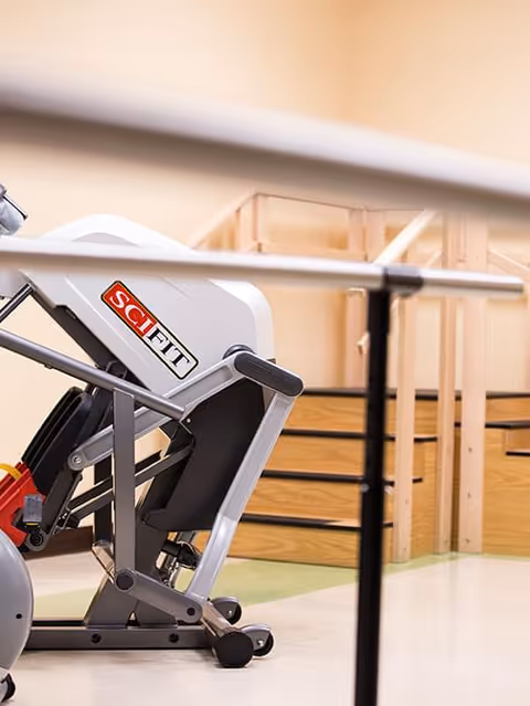 A close-up view of a SciFit exercise machine in a rehabilitation or fitness room with wooden handrails and steps in the background.