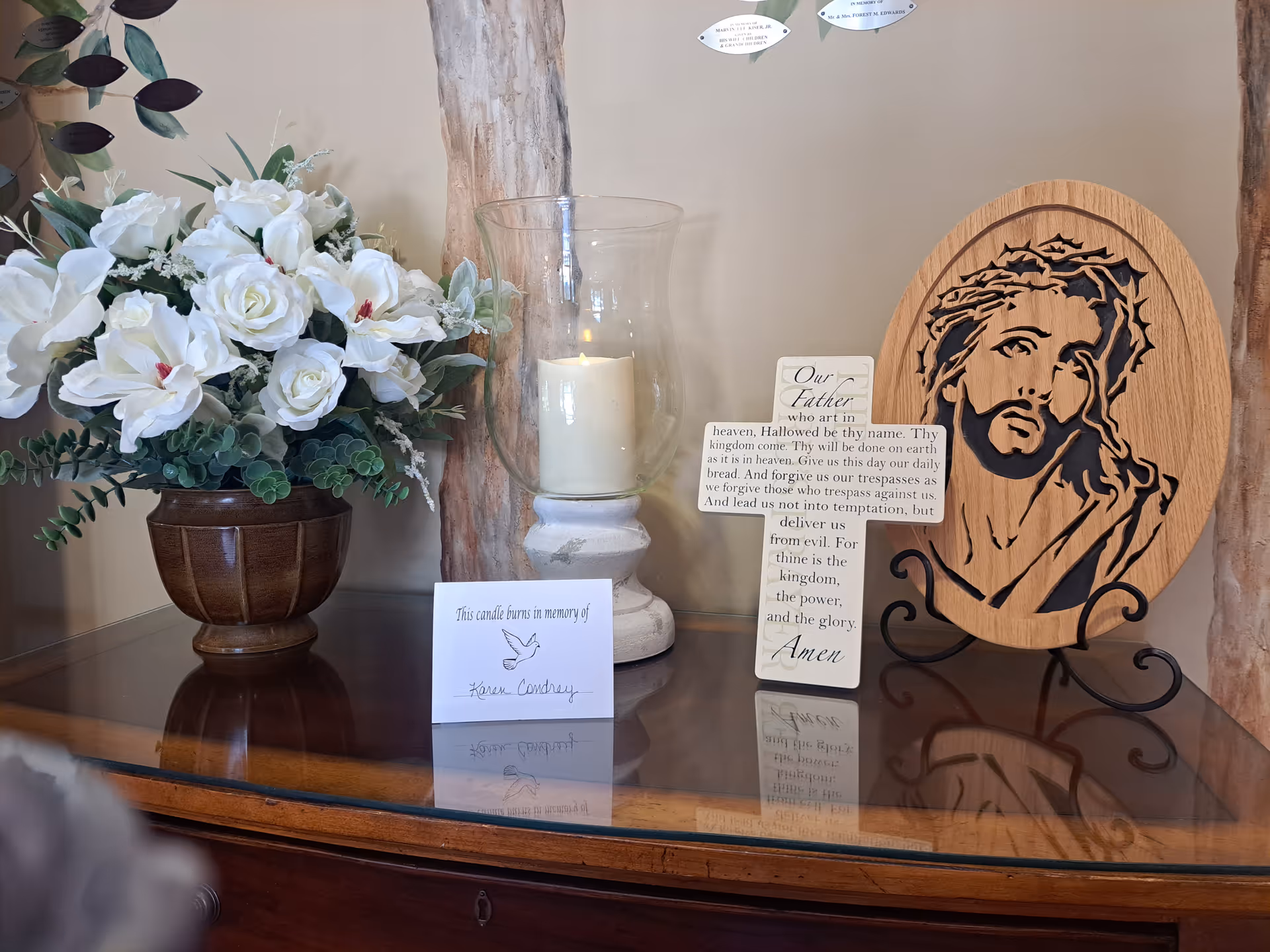 Floral bouquet, lit candle, and wooden Jesus plaque with memorial cards on a polished table.