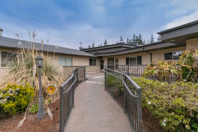 Concrete ramp with metal railings leading to the entrance of a single-story rehabilitation facility, flanked by shrubs and ornamental grasses.