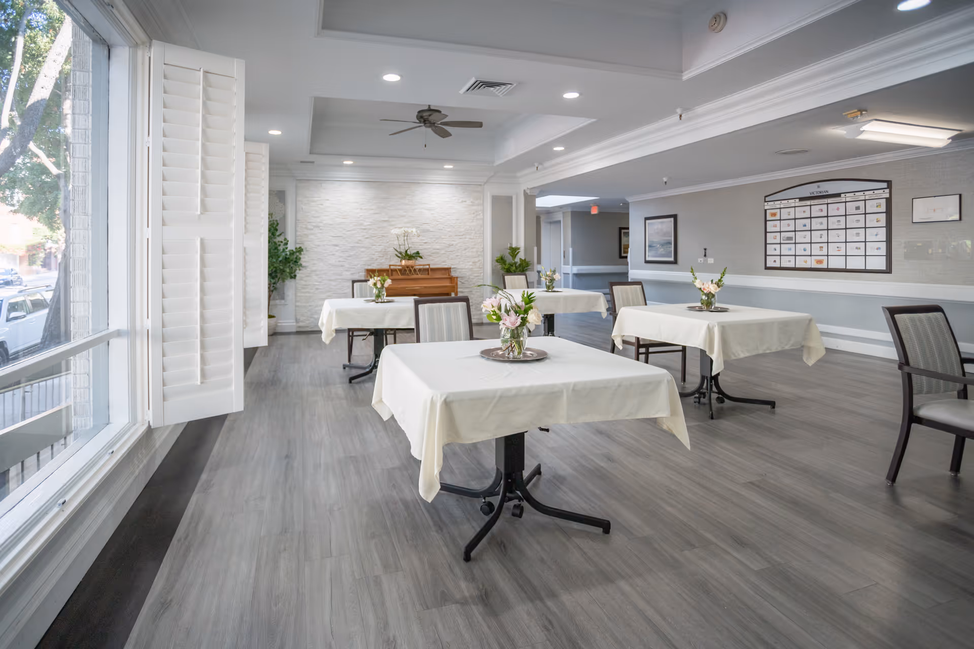 Bright dining room with white-clothed tables, chairs, flowers, and a piano in back.