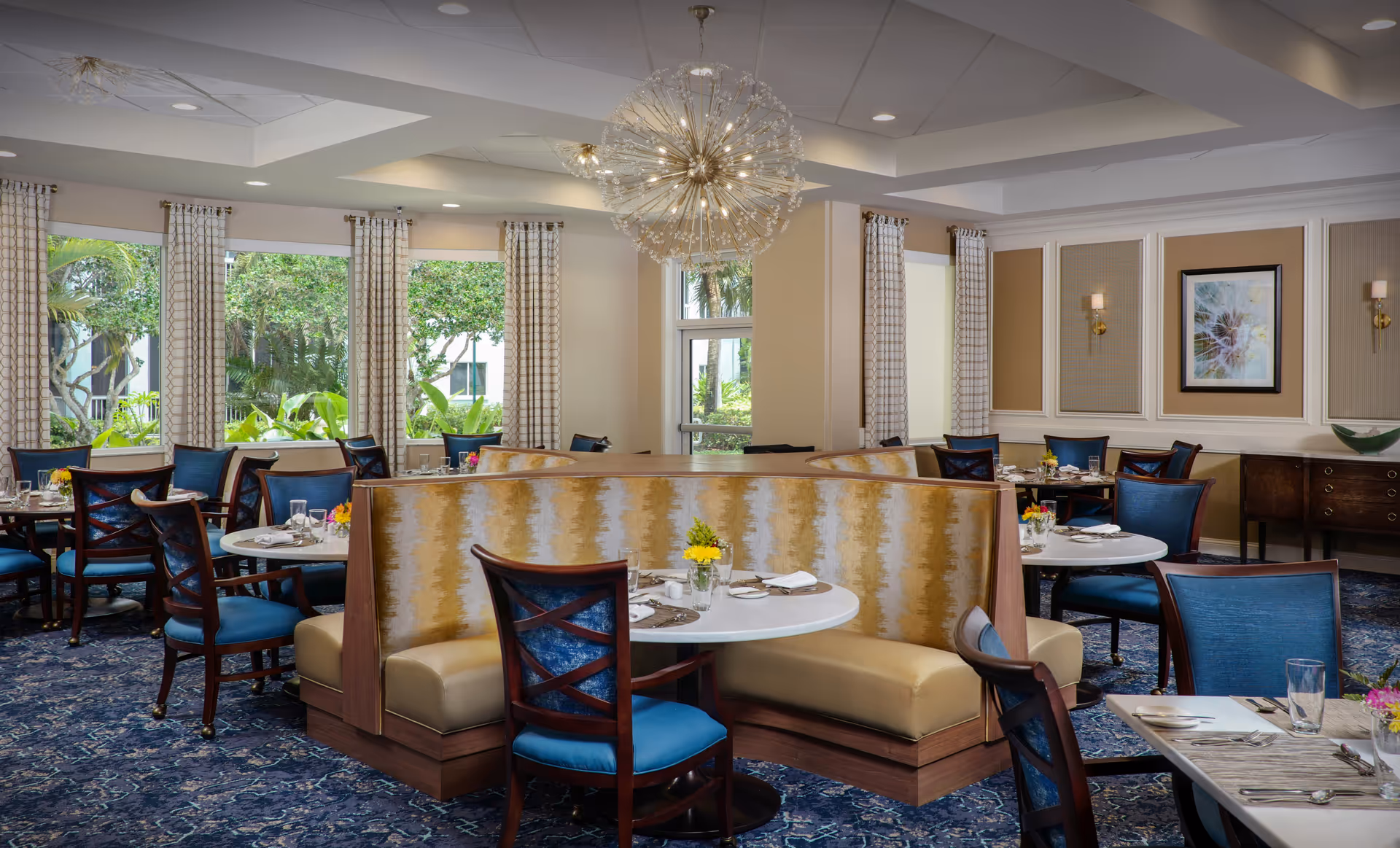 Elegant dining room with round tables, blue chairs, beige booths, a chandelier, and large windows.