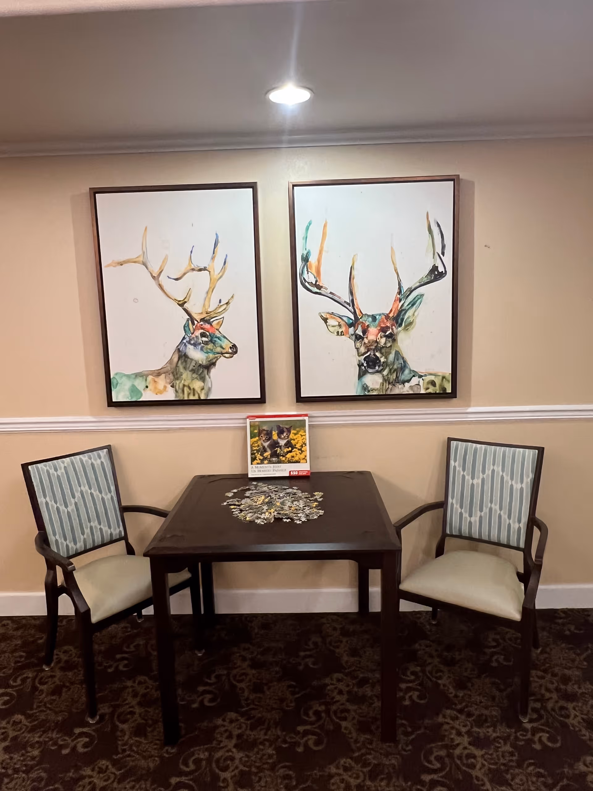 A small dark wooden table with a partially completed jigsaw puzzle and a puzzle box on top, flanked by two wooden chairs with light blue patterned cushions. Above the table are two framed colorful watercolor paintings of deer heads mounted on a beige wall with white trim.