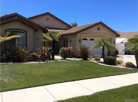 Single-story residential building with a tiled roof, a garage, and a well-maintained front yard featuring green grass, shrubs, and small palm trees under a clear blue sky.