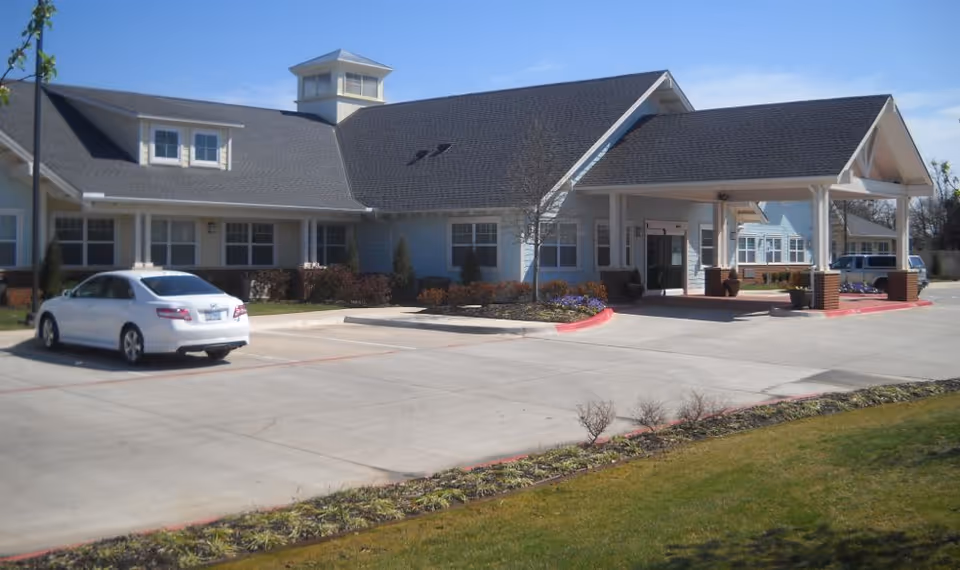 Exterior view of a single-story senior living facility building with a covered entrance, multiple windows, a small landscaped area with shrubs and flowers, and a white car parked in the parking lot on a sunny day.