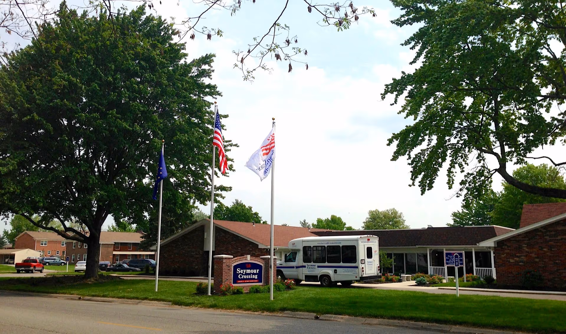 Exterior front of Seymour Crossing senior living facility showing the entrance sign, flags, and a shuttle bus.