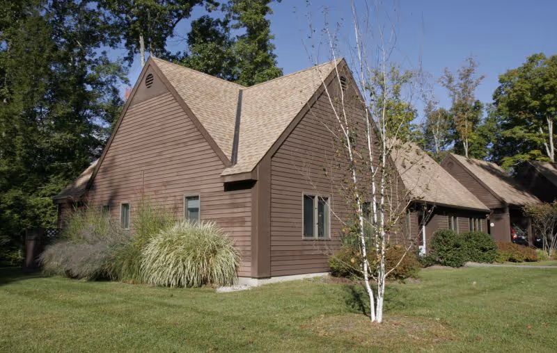 Brown wood-clad single-story building with gabled roofs, lawn, and trees in front.
