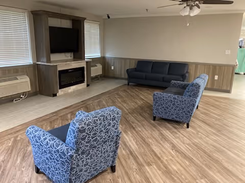 A senior living facility common area with two patterned blue armchairs and a dark blue sofa arranged on a wood-patterned floor. There is a wall-mounted TV above an electric fireplace, two windows with blinds, and ceiling fan with lights. The walls are painted beige with wood paneling on the lower half.