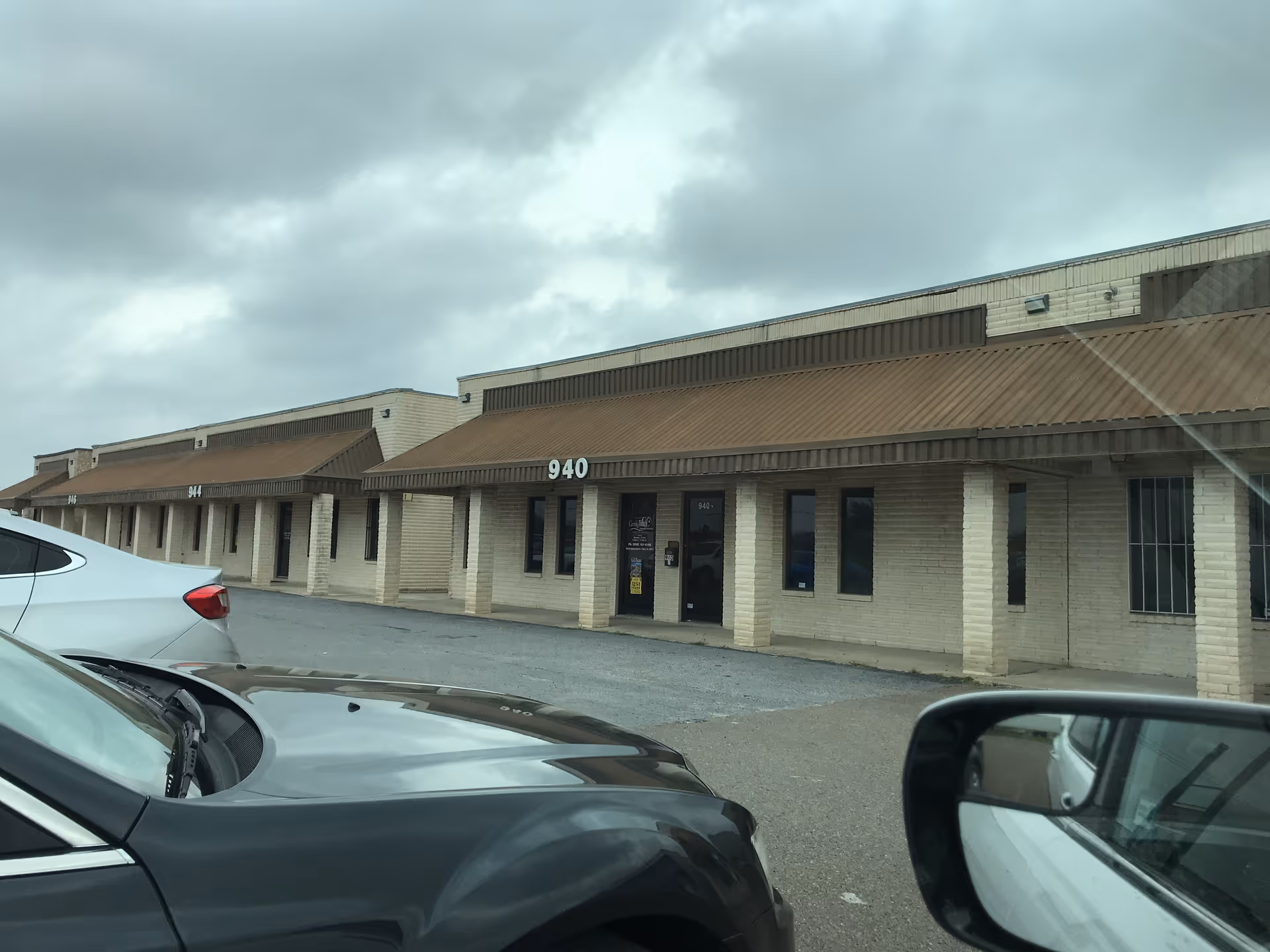 Row of beige brick storefronts with brown awnings and parked cars in front.