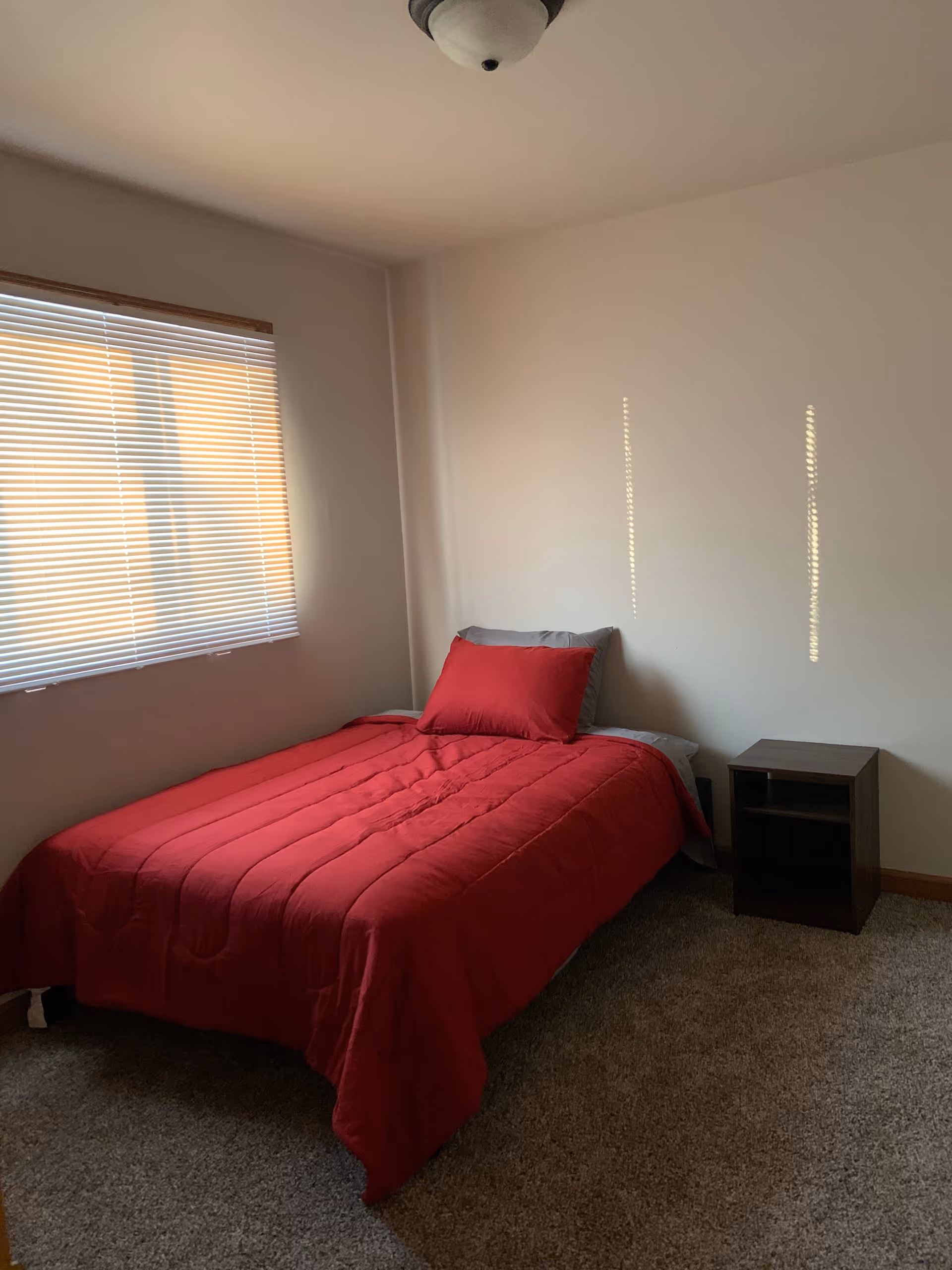 A small bedroom with a red bedspread, window blinds, and a nightstand.