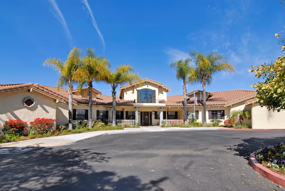 Exterior view of a single-story senior living facility with palm trees and a parking area.